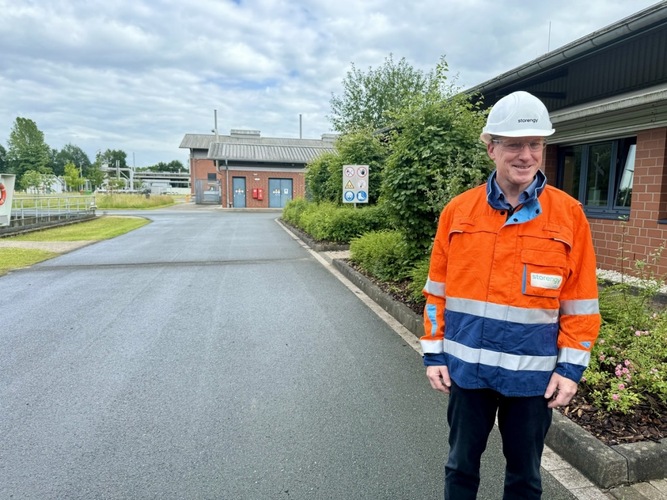 Man in orange safety jacket and white hard hat stands on paved road near industrial buildings and greenery. Man in orange safety jacket and white hard hat stands on paved road near industrial buildings and greenery.